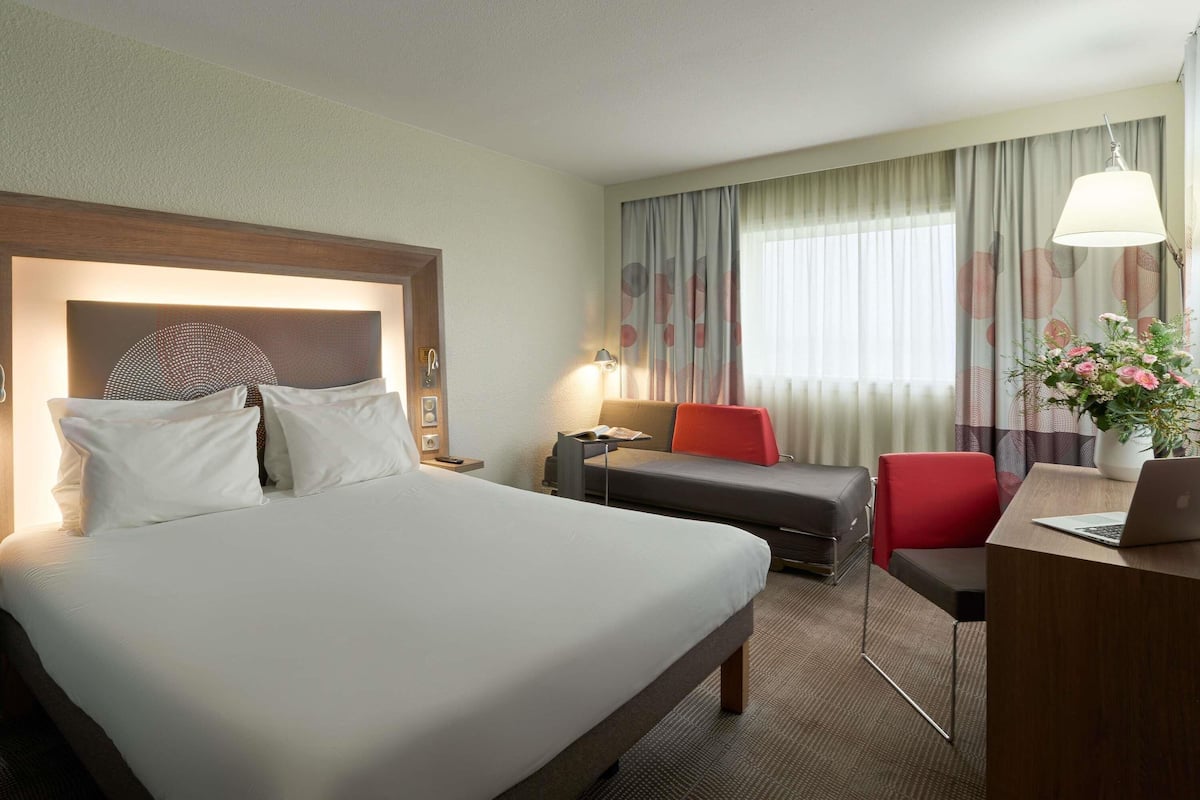 A hotel room with a large bed, a desk with a laptop, a chair, a sofa, and a window with curtains.