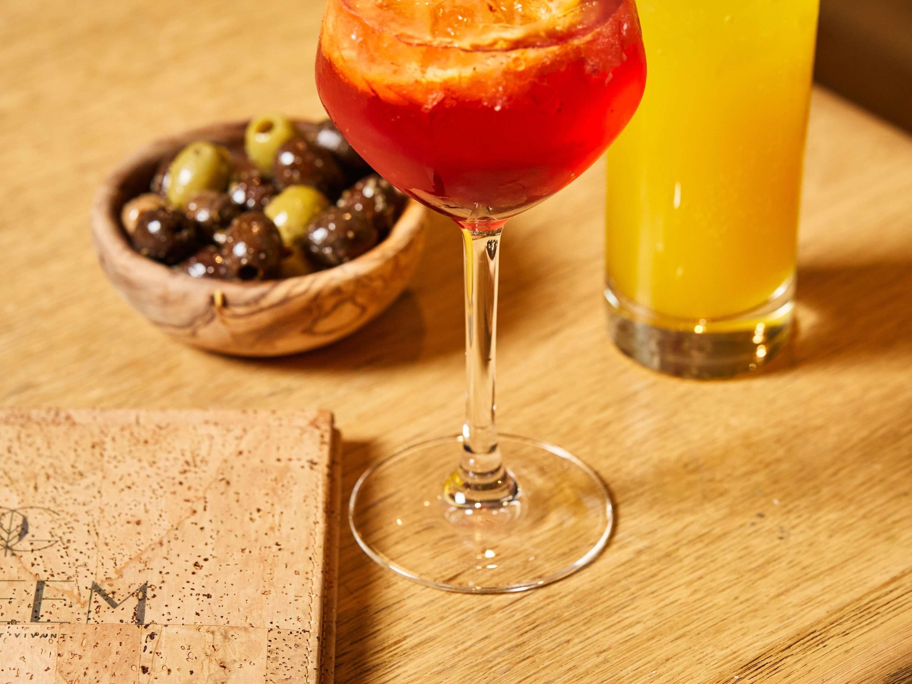Un verre à cocktail contenant une boisson rouge, un verre de jus d’orange et un bol d’olives sur une table en bois.