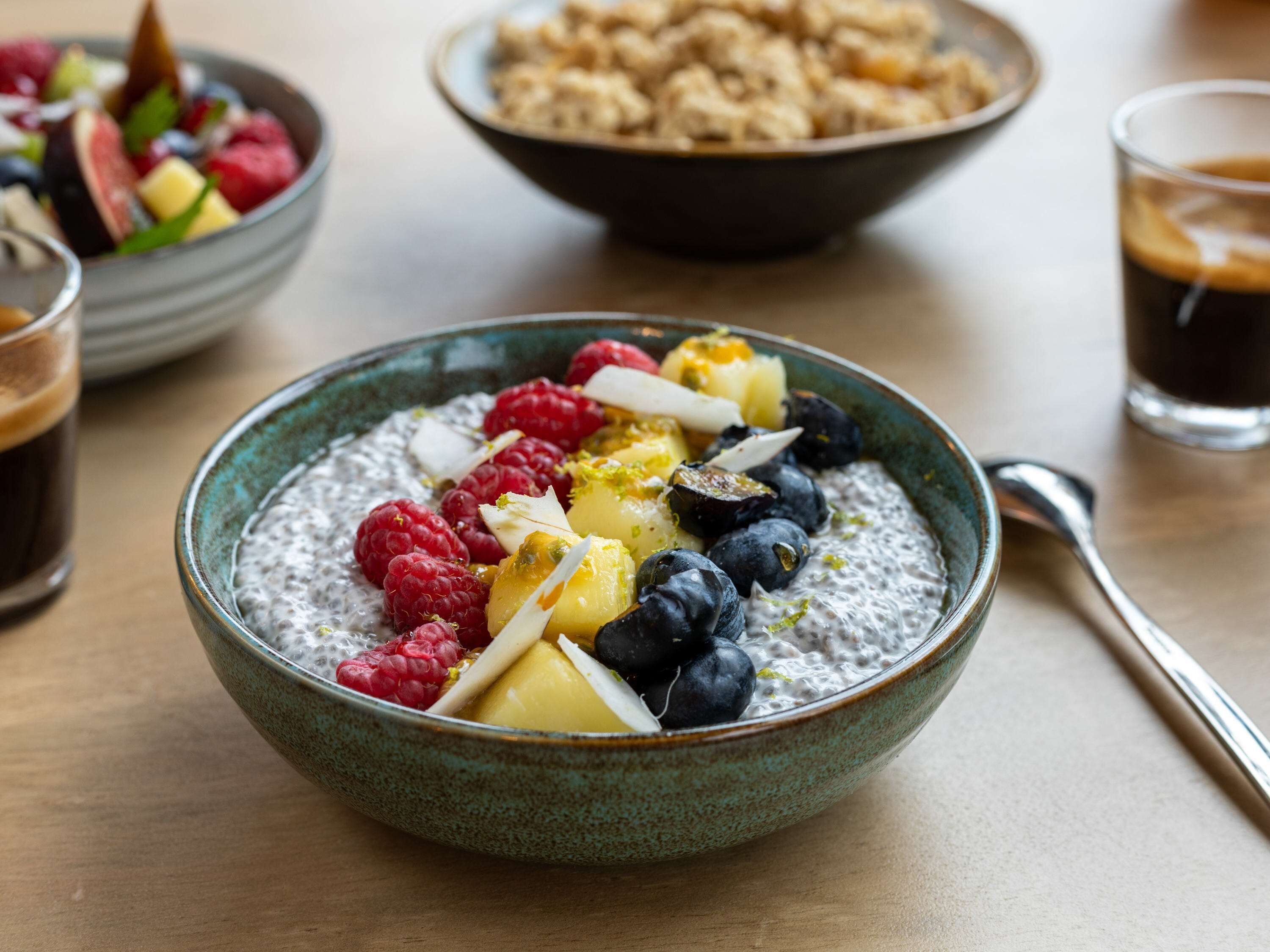 A bowl of oatmeal topped with fruits and nuts, a glass of coffee, and a bowl of granola in the background.