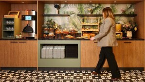 A woman walking past a counter with baked goods and a coffee machine. - Novotel Paris La Défense Esplanade (Courbevoie)
