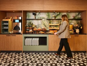 A woman walking past a counter with baked goods and a coffee machine. - Novotel Paris La Défense Esplanade (Courbevoie)