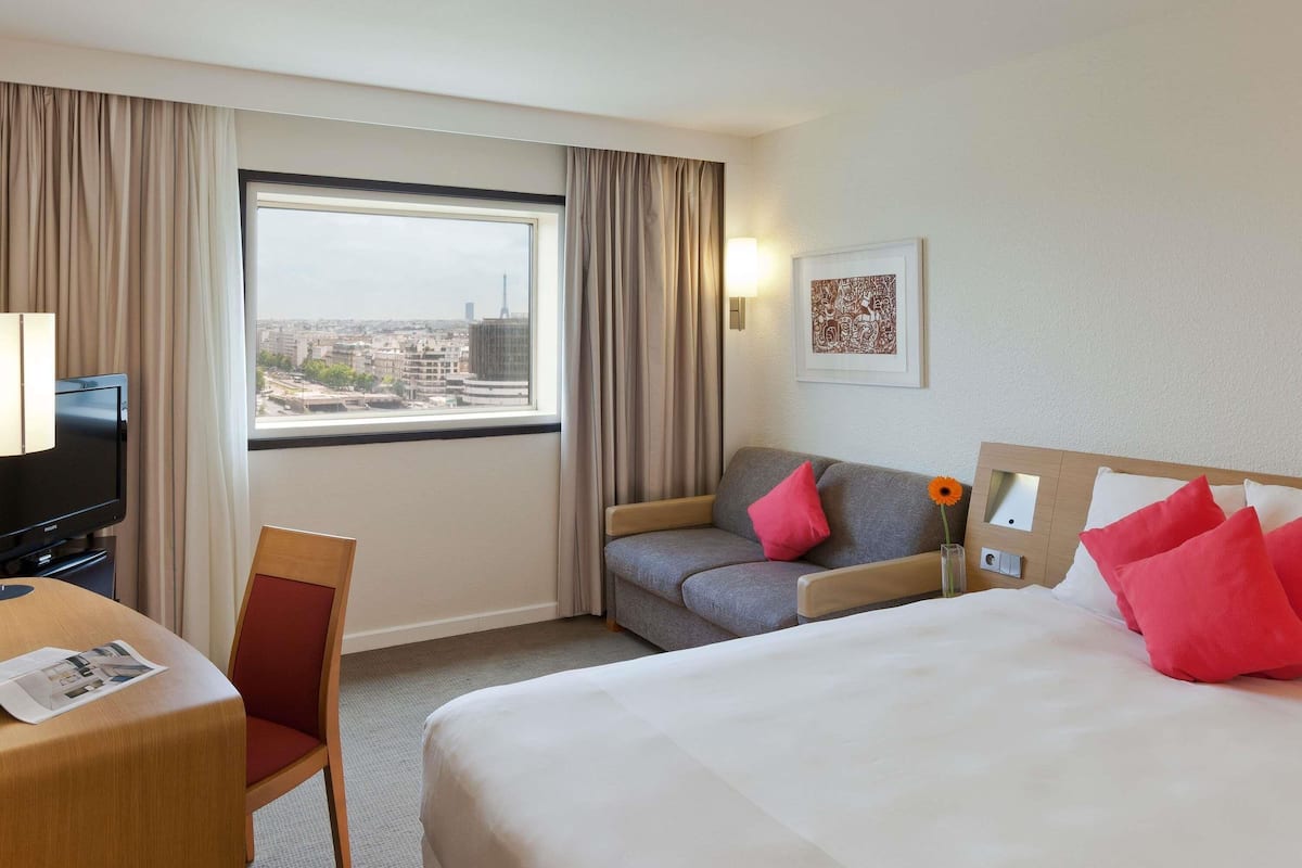 A hotel room with a bed, a sofa, a TV, a desk, and a window with a city view.