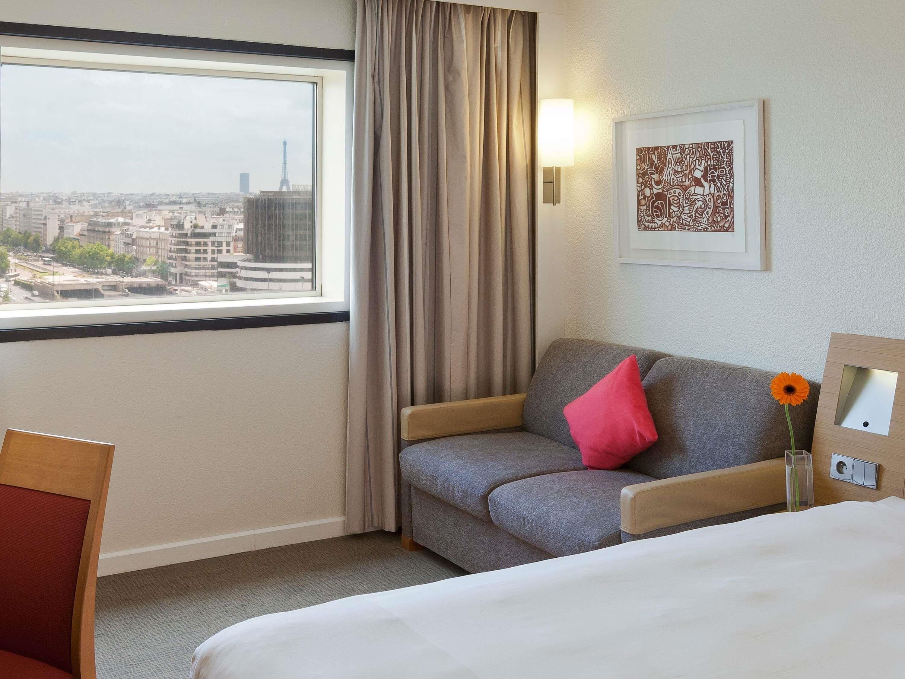 Ein Hotelzimmer mit einem Bett, einem grauen Sofa mit rotem Kissen, einem Fenster mit Blick auf Gebäude, einer Lampe und einem Bilderrahmen an der Wand.