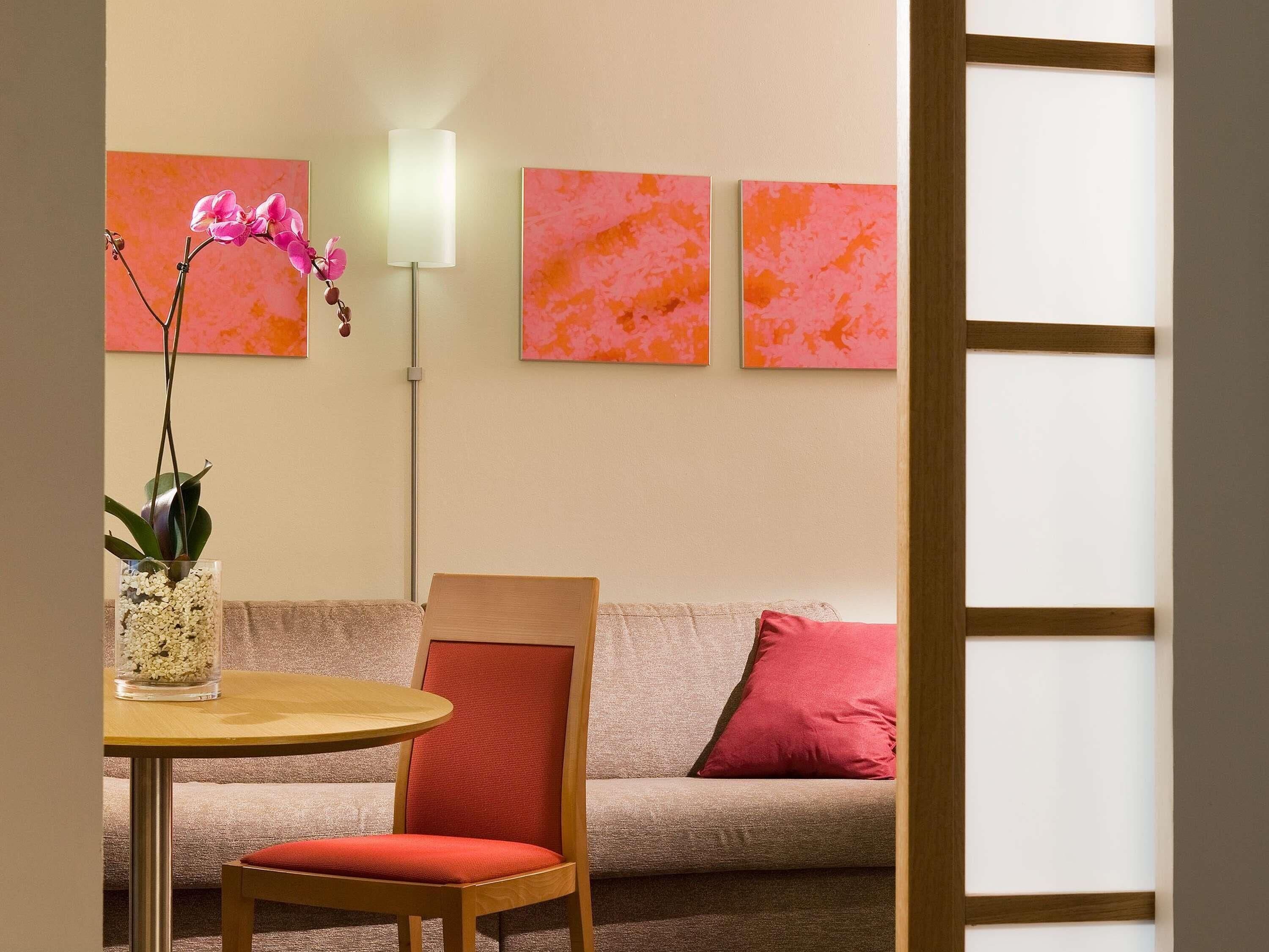 A room with a sofa, a chair, a table, a vase with flowers, and three paintings on the wall.