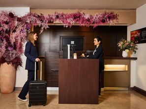 A reception desk with a dark wood finish, a large potted plant to the left, and a wall-mounted television above the desk. - Novotel Paris La Défense Esplanade (Courbevoie)