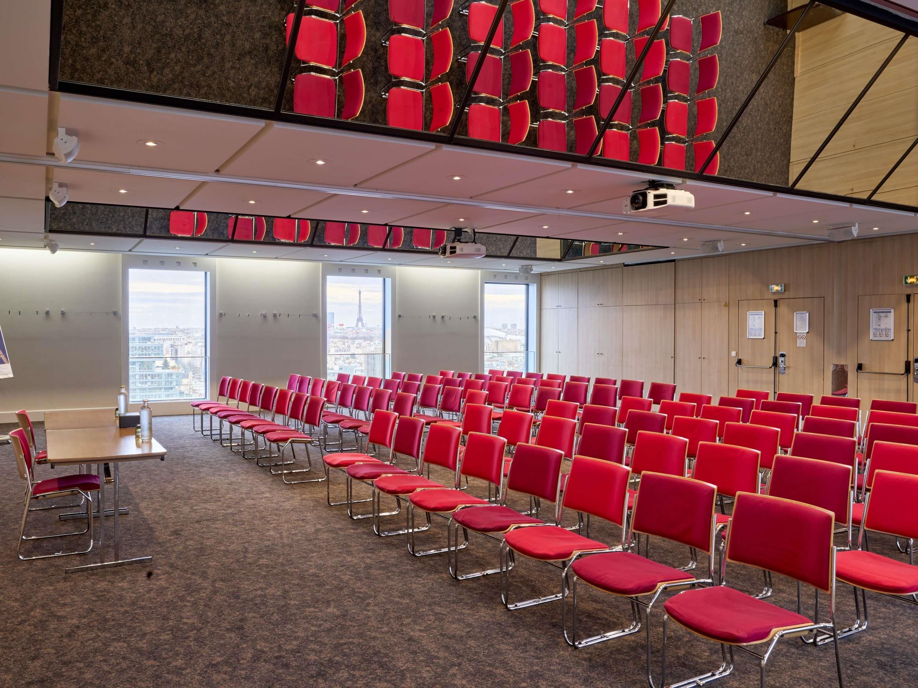 A conference room with rows of red chairs, a projector, and a large window with a city view.