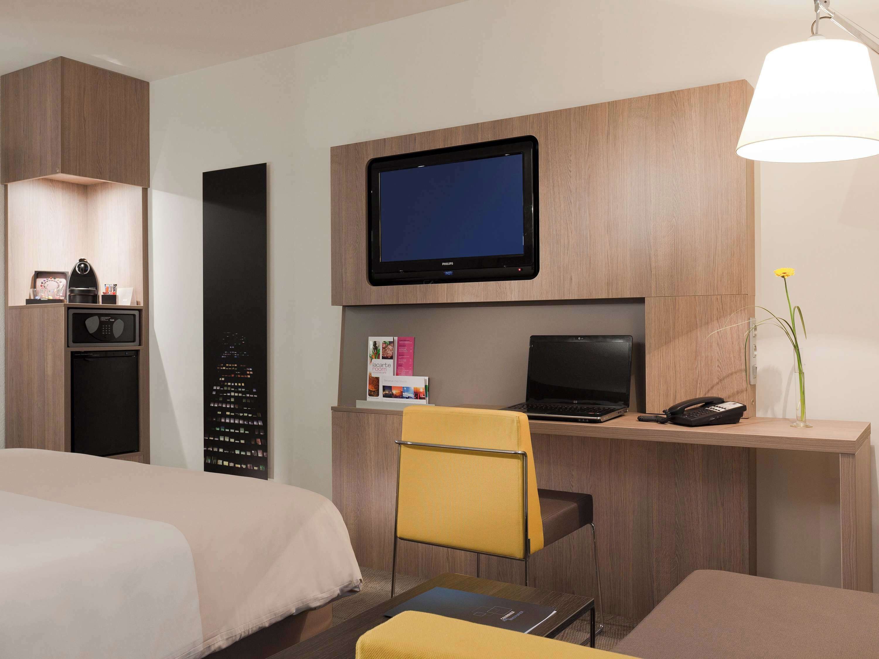 Una camera d'albergo con un letto, una scrivania con un portatile, una televisione, un telefono e un piccolo vaso con un fiore.