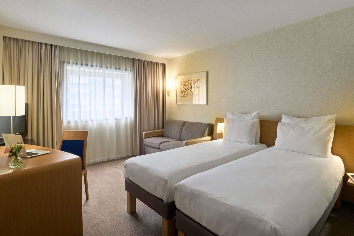 A hotel room with two beds, a desk with a laptop, a chair, and a large window with curtains.