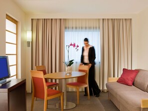 Quarto de hotel com mesa de jantar, cadeiras, sofá, televisão e janela com cortinas.
