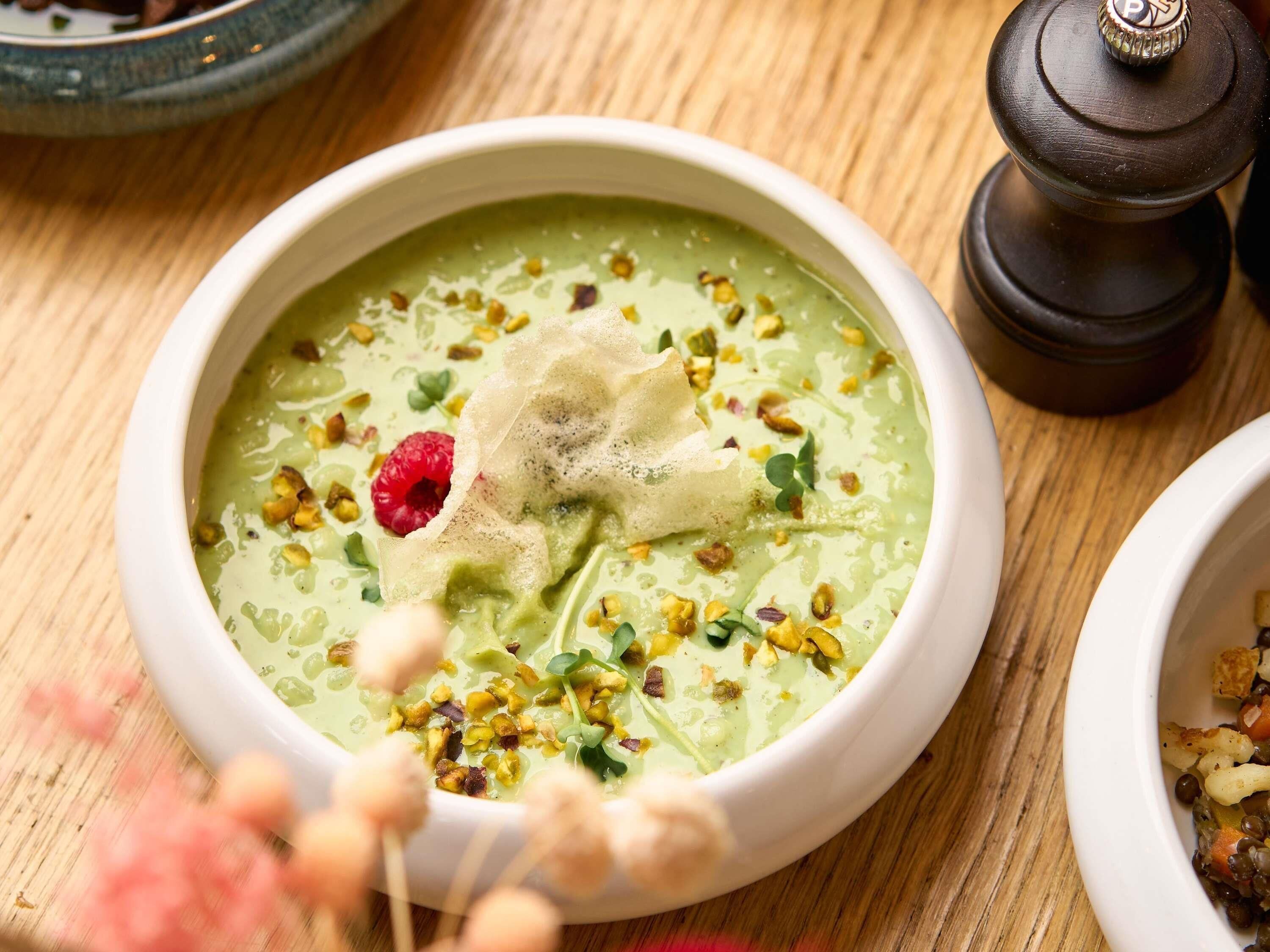 Un bol de soupe verte, garni de noix et d’une framboise, avec un moulin à poivre et une table en bois en arrière-plan.