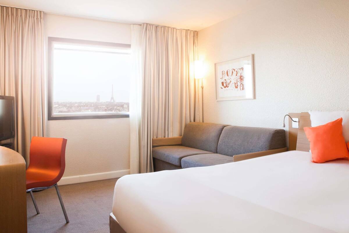 A hotel room with a bed, a sofa, a chair, a TV, and a window with a city view.