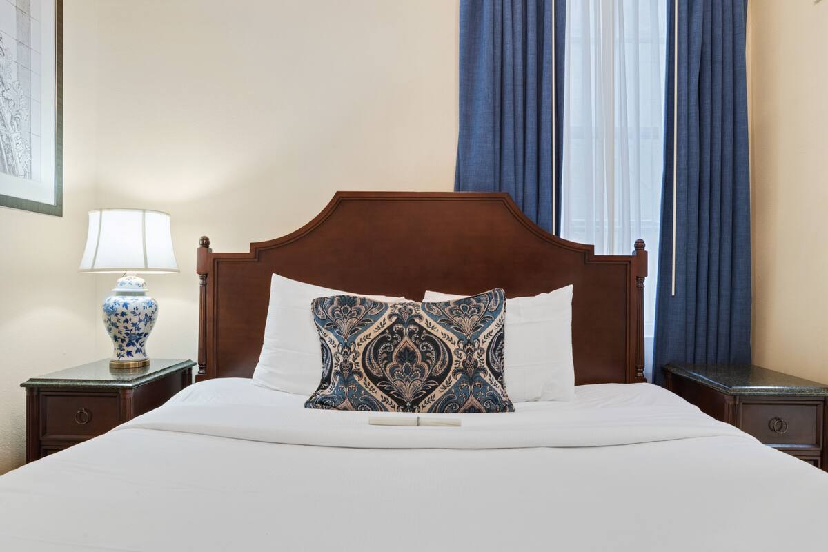 A neatly made bed with a wooden headboard, a decorative pillow, and a nightstand with a lamp and a vase.