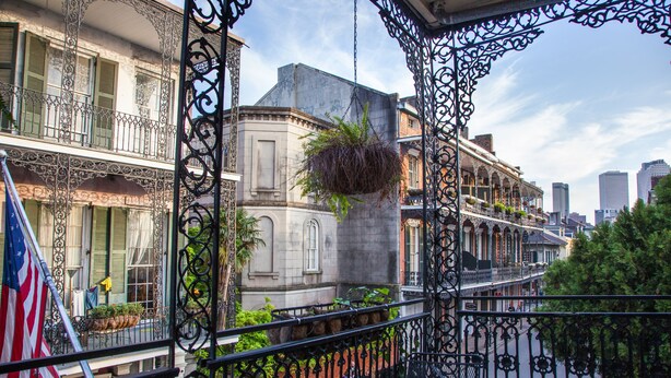 Un balcon orné de balustrades en fer forgé, une plante suspendue et une vue sur un paysage urbain avec des bâtiments et un drapeau.