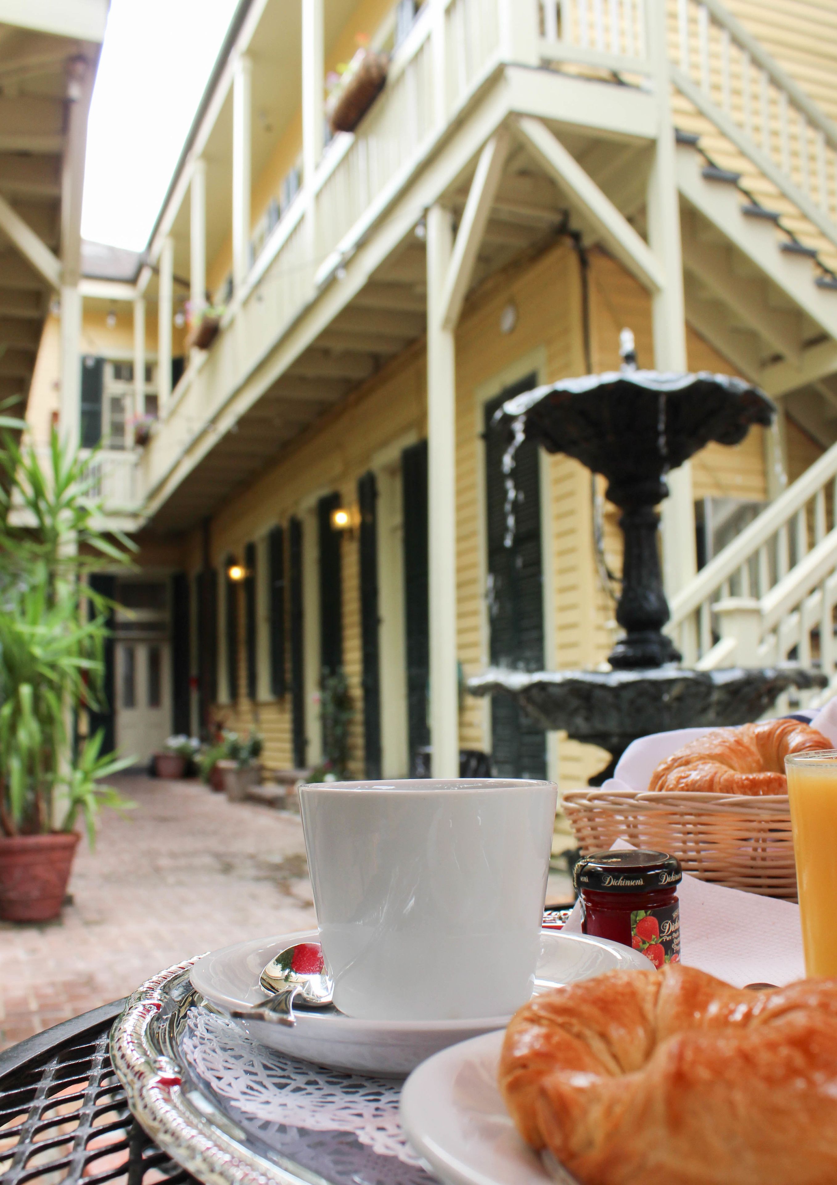 A table set with a cup, a croissant, and a bottle of jam, with a fountain and a building in the background.