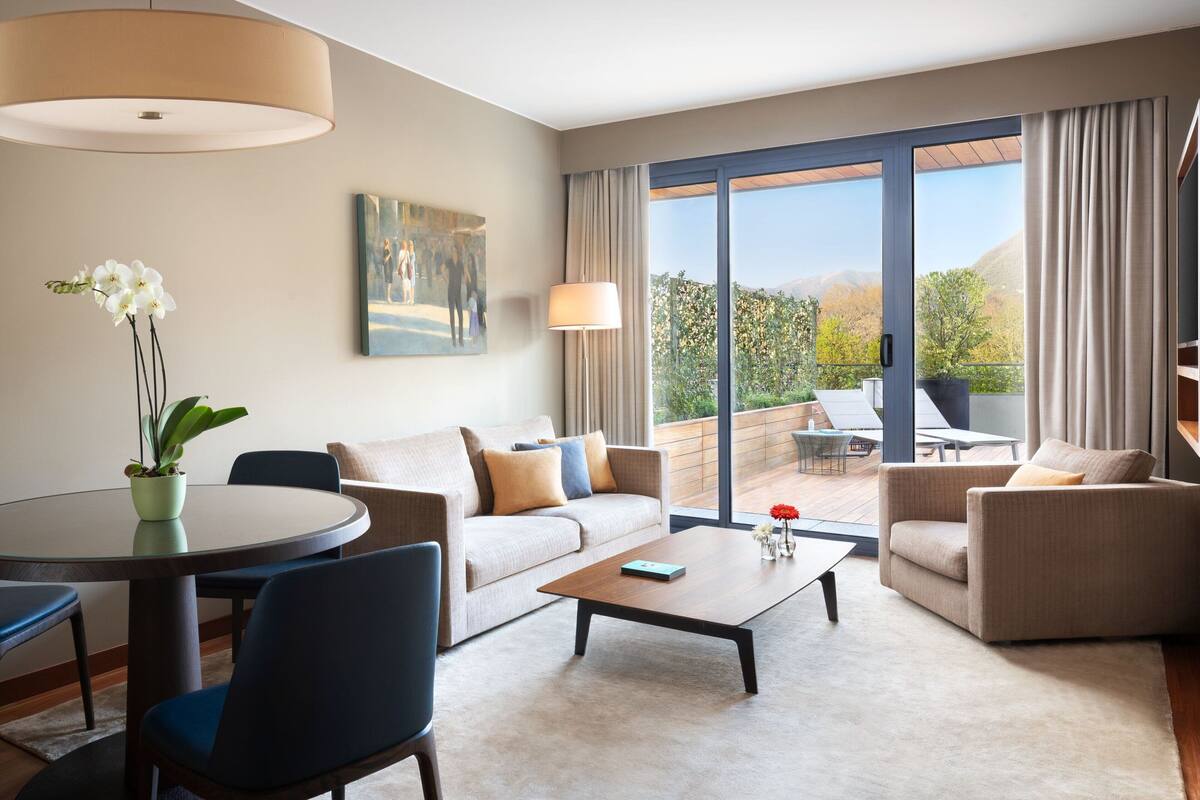 A modern hotel room with a large sliding glass door to a balcony, a round dining table, a sofa, and a coffee table.