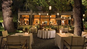 An outdoor dining area with tables set for two, surrounded by trees and a building with large windows. - Sheraton Lake Como Hotel (Como)