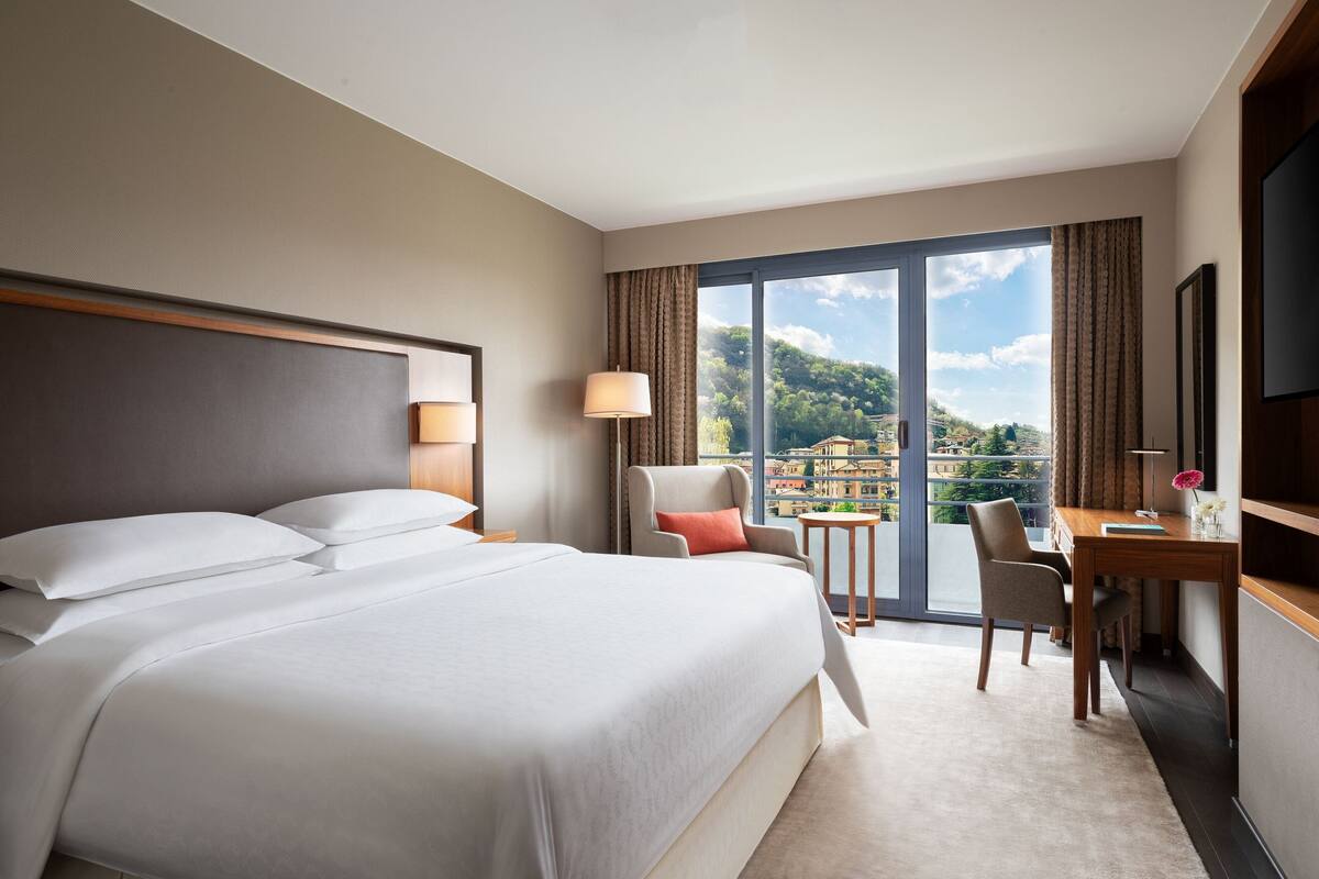 A hotel room with a large bed, a desk, a chair, and a view of a cityscape through large windows.