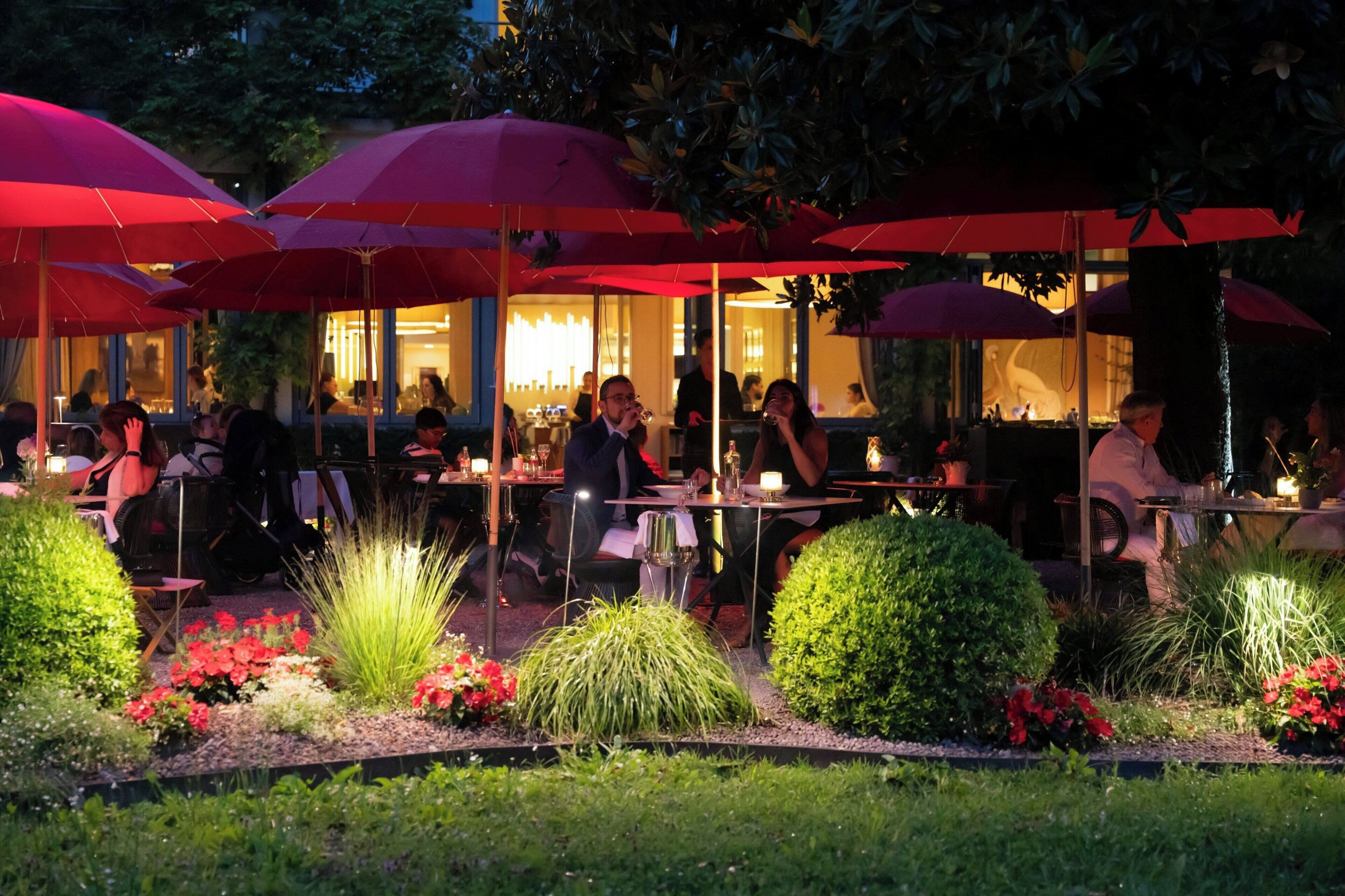 An outdoor dining area with tables and chairs, surrounded by red umbrellas and greenery.