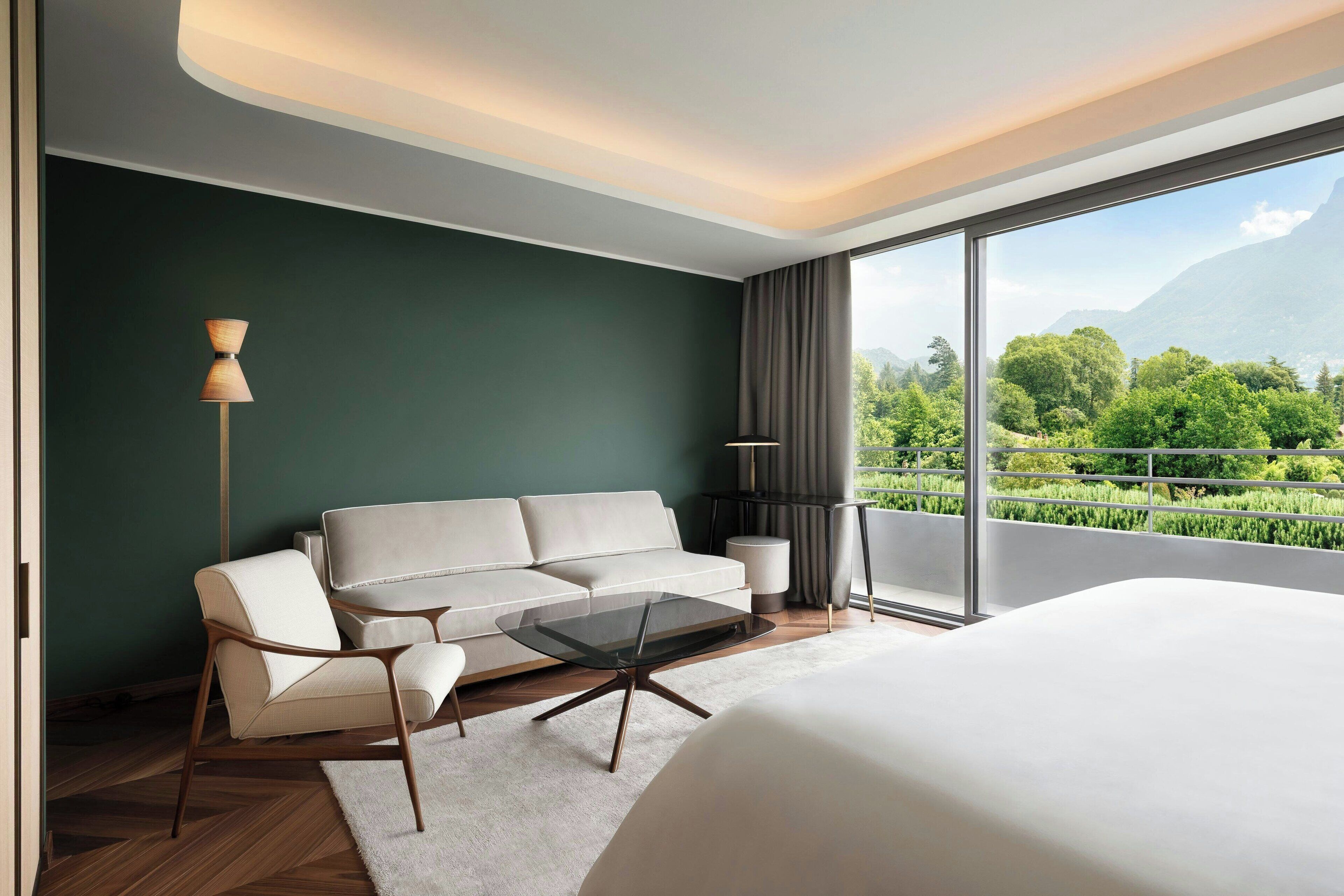 Habitación de hotel moderna con una cama grande, un sofá, una mesa de centro de vidrio y vistas a un paisaje verde exuberante a través de grandes ventanales.