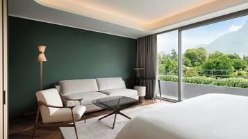 Habitación de hotel moderna con una cama grande, un sofá, una mesa de centro de vidrio y vistas a un paisaje verde exuberante a través de grandes ventanales.