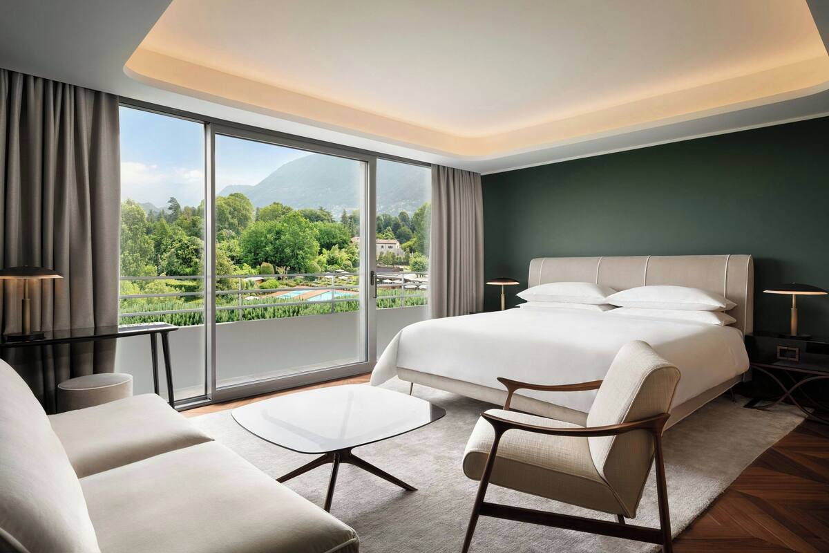 A modern hotel room with a large bed, a sitting area with a table, and a view of greenery and mountains through large windows.