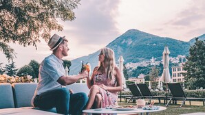 A couple sitting on a sofa outdoors with a table between them, overlooking a mountainous landscape and a town in the background. - Sheraton Lake Como Hotel (Como)