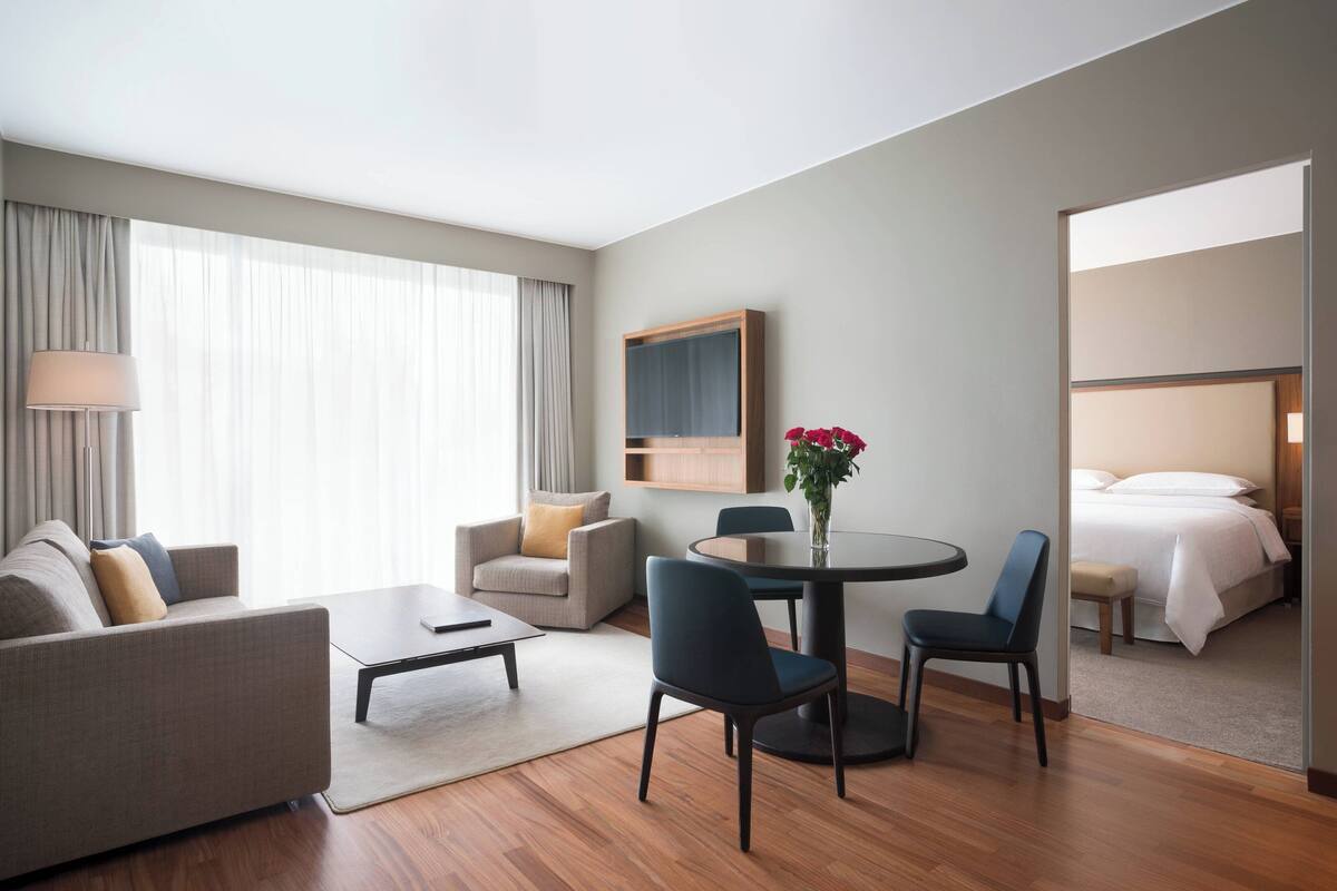A modern hotel room with a sofa, two armchairs, a small table, a bed, a mirror, and a window with curtains.
