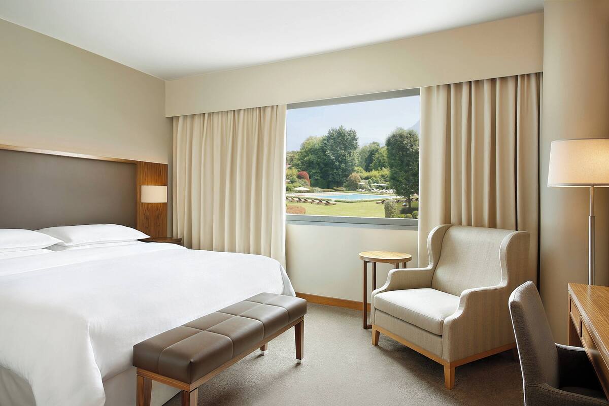 A hotel room with a large bed, a bench, two armchairs, a small table, and a view of a garden through a window.