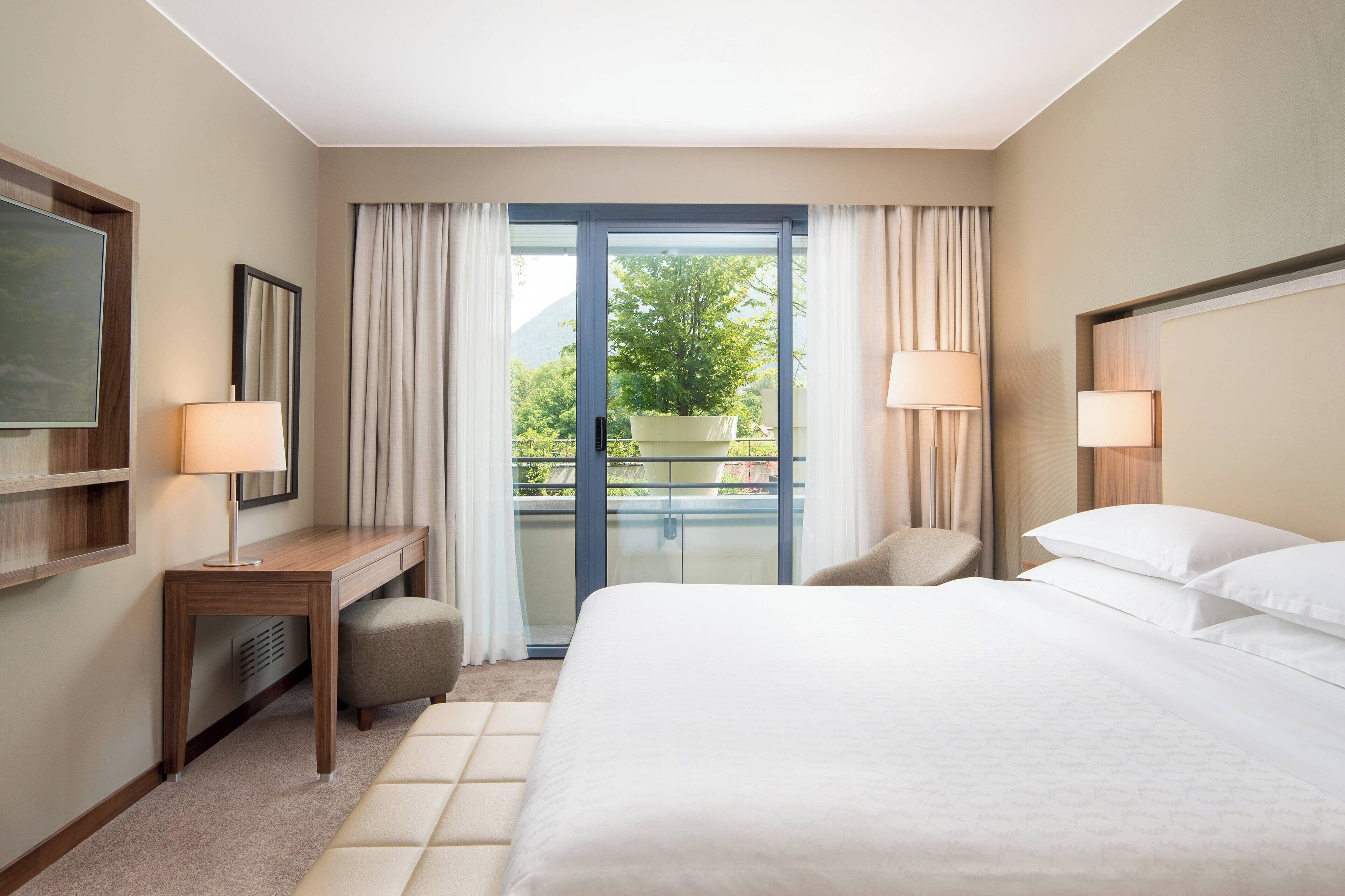 Camera d'albergo con letto, scrivania, sedia, televisore e balcone con vista sul verde.
