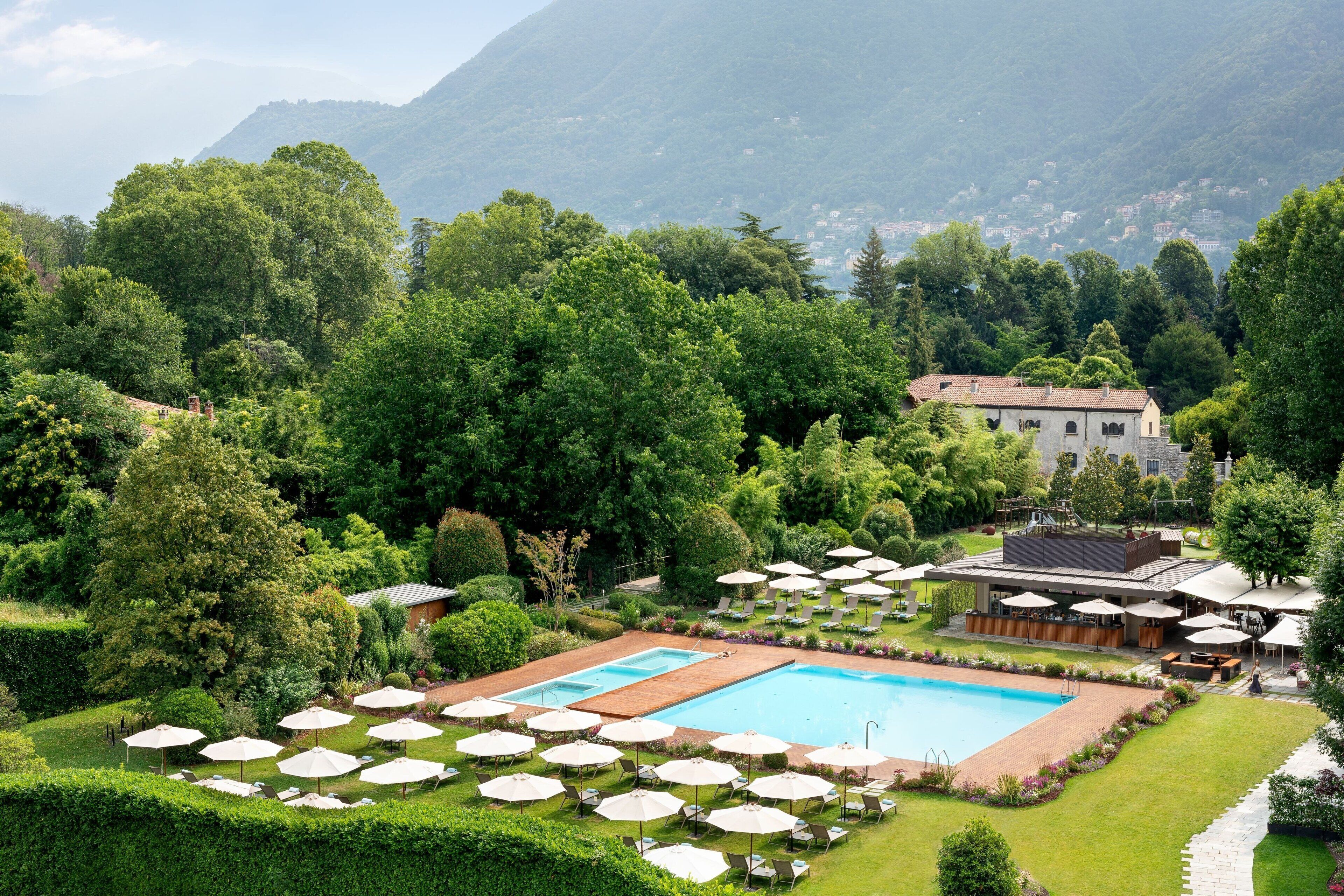 Ein Poolbereich mit mehreren Liegestühlen und Sonnenschirmen, umgeben von gepflegter Bepflanzung und einem Bergpanorama.