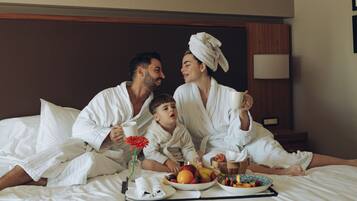 Une famille de quatre personnes dans une chambre d’hôtel, assise sur le lit, avec un plateau de petit-déjeuner comprenant des fruits et une fleur.