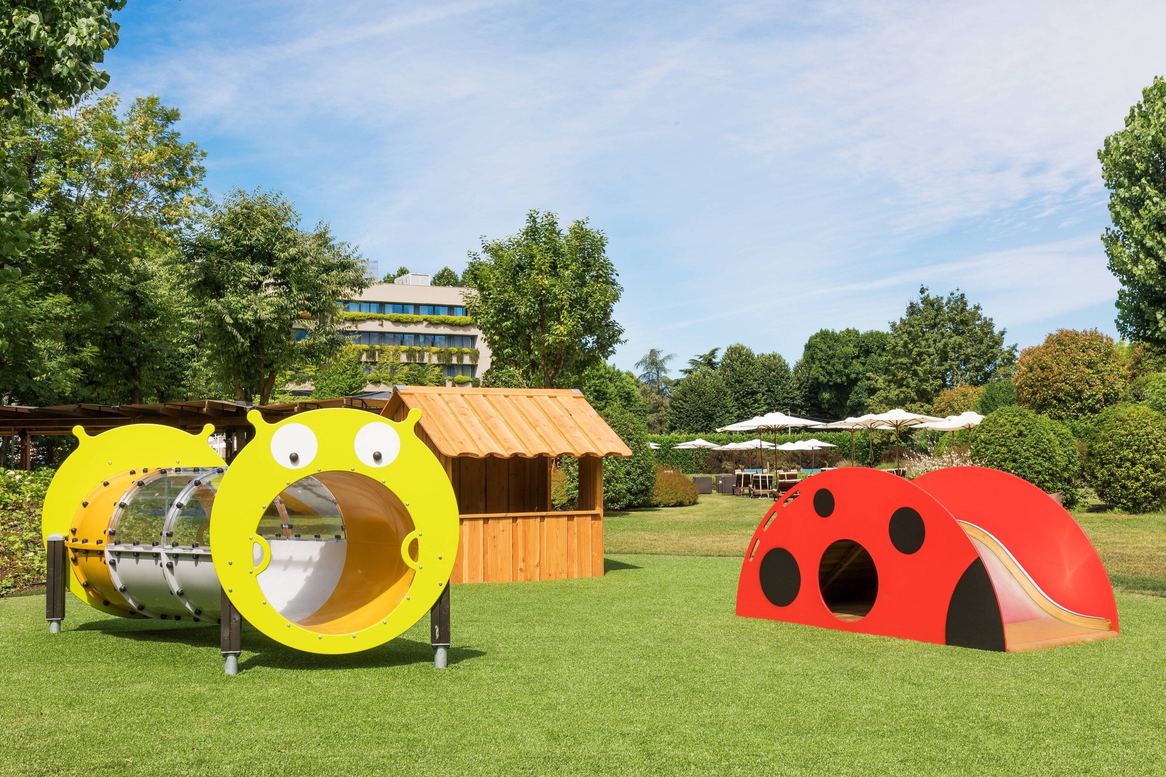 A playground with slides and tunnels, a wooden playhouse, and a building in the background.