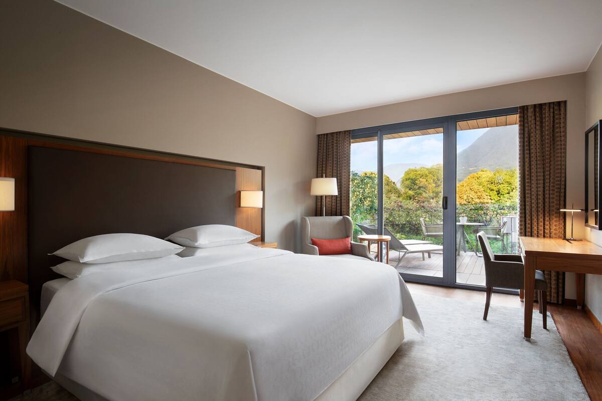 A hotel room with a large bed, a desk, and a chair. There is a balcony with a table and chairs.