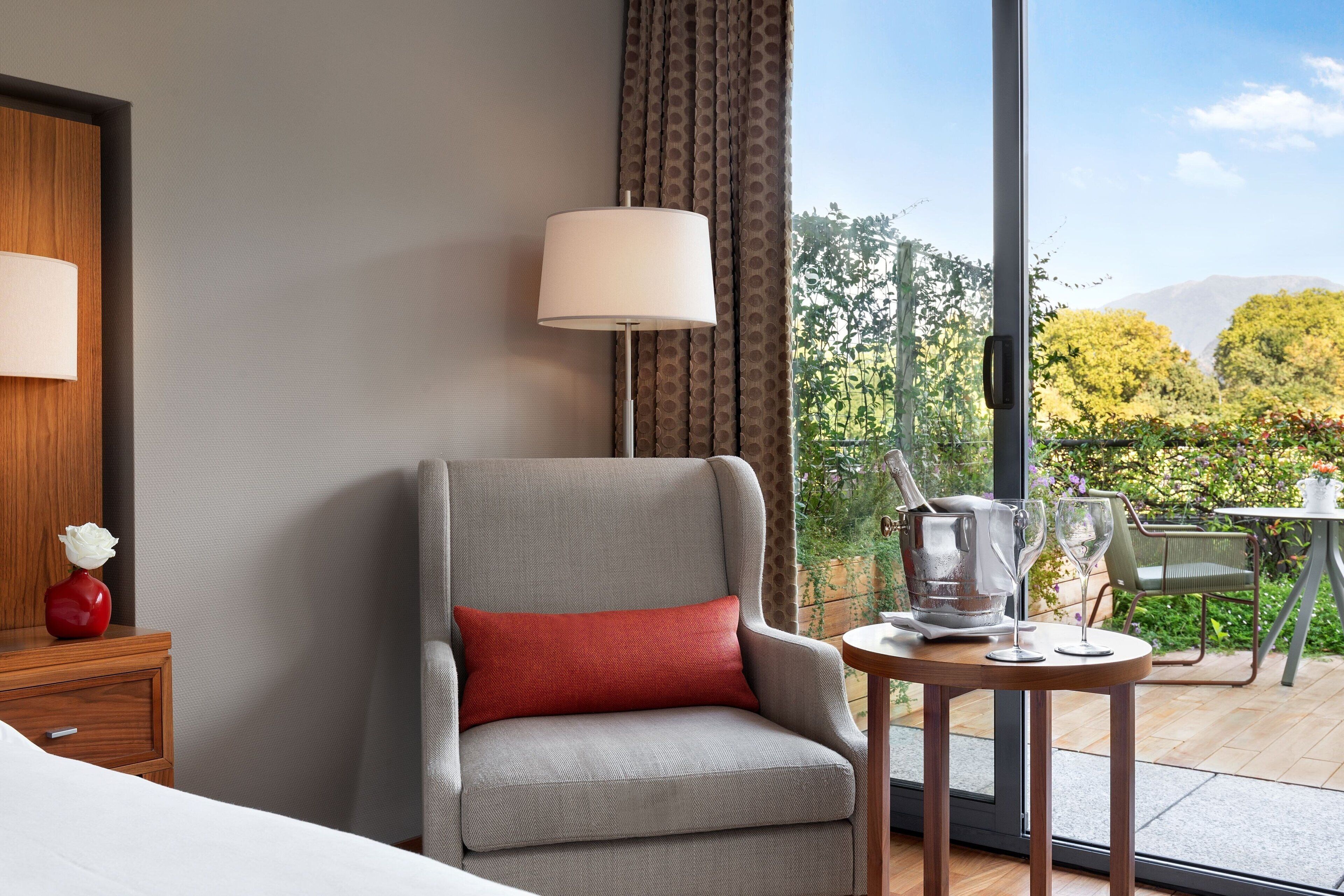 Habitación de hotel con un sillón gris, una pequeña mesa con una jarra y vasos, una mesita de noche de madera con un florero, y un ventanal que ofrece vistas a la vegetación y montañas.
