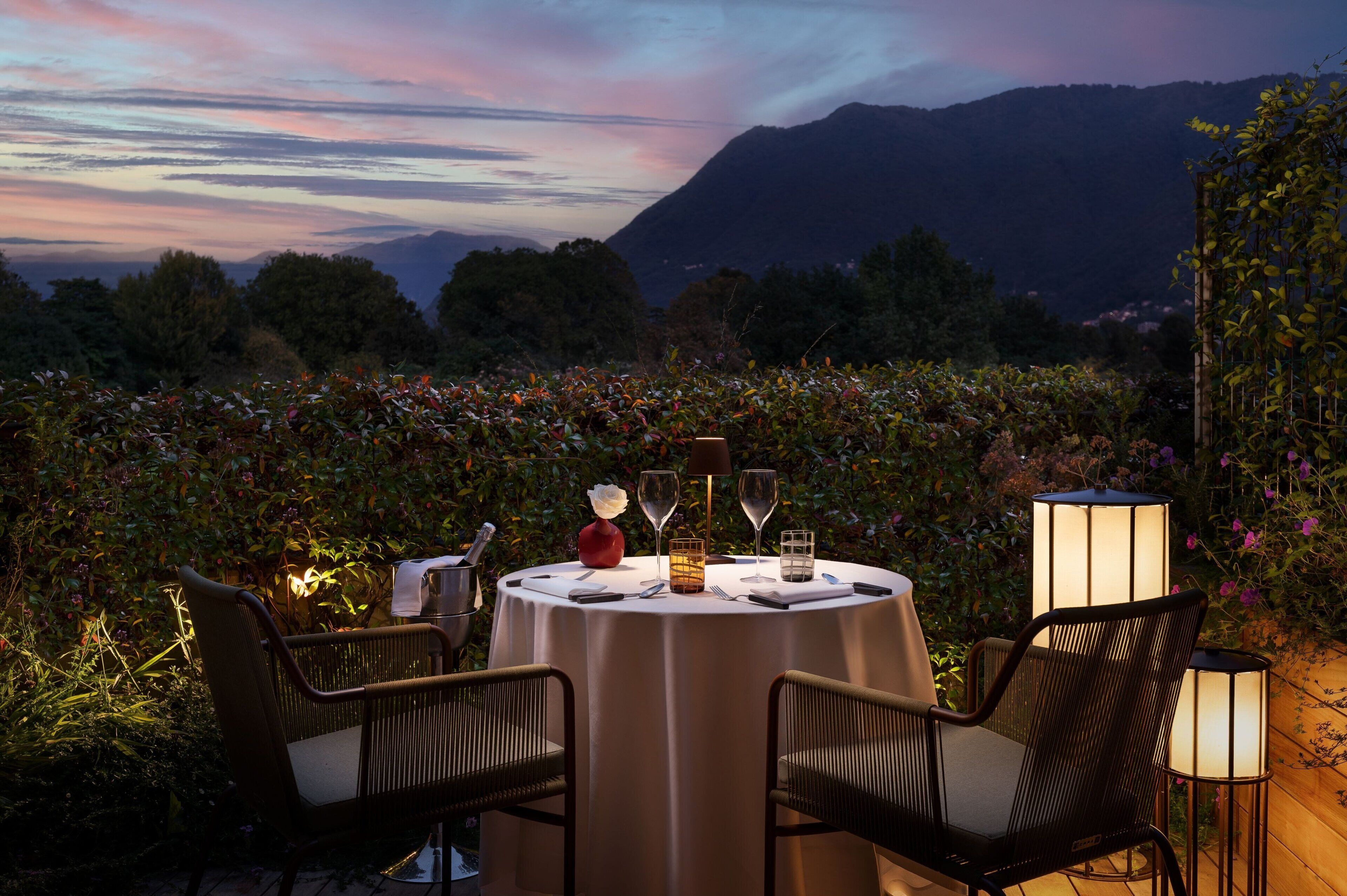 Un área de comedor al aire libre con una mesa redonda preparada para dos personas, rodeada de sillas, con montañas al fondo y exuberante vegetación.