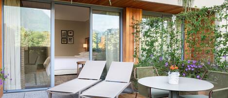 Un espacio lounge exterior moderno con terraza de madera, sillas de descanso blancas, una mesa redonda y vista a un dormitorio a través de puertas de vidrio.