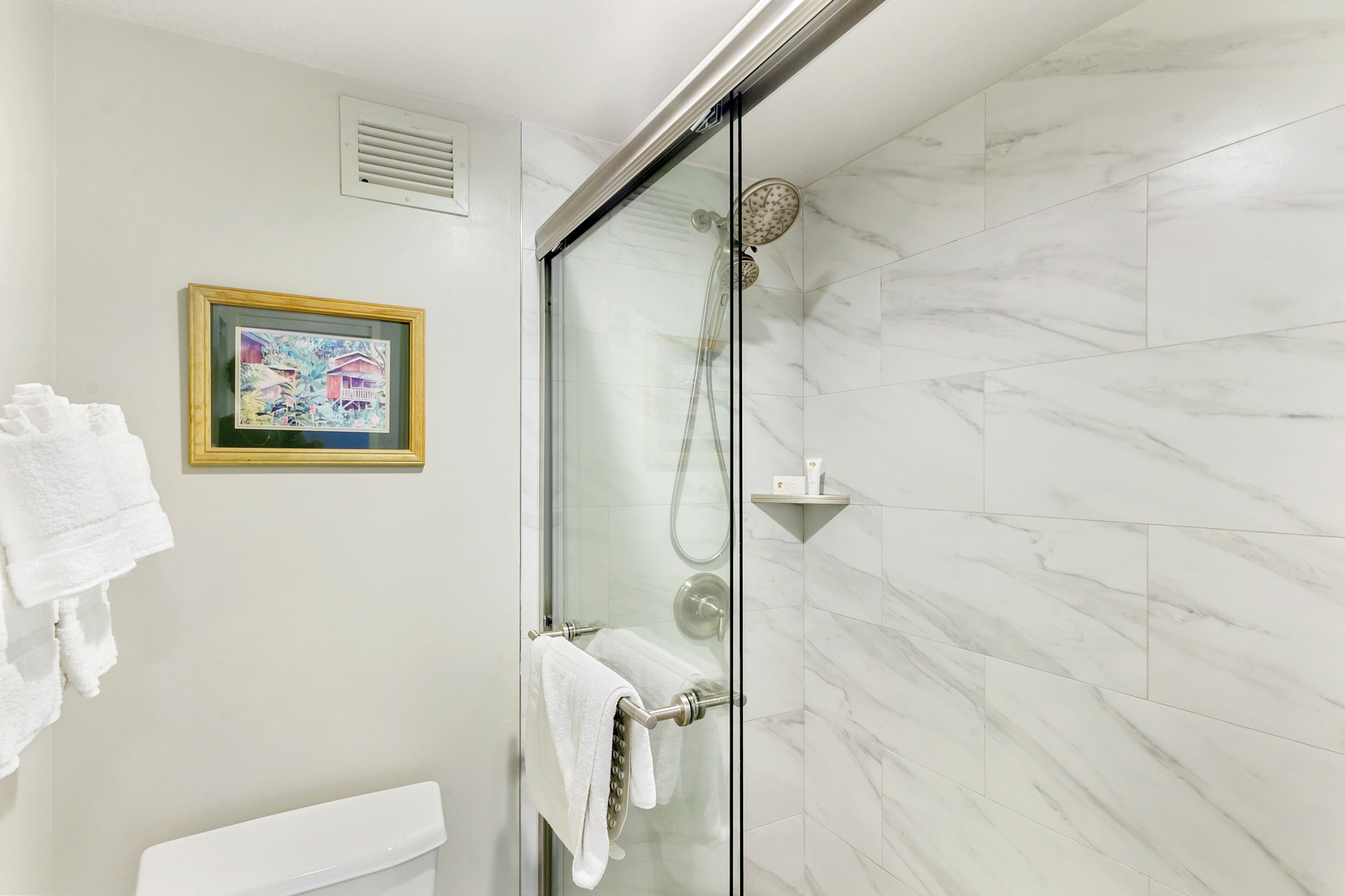Un baño moderno con ducha envuelta en vidrio, baldosas de mármol blanco, un cuadro enmarcado en la pared y un perchero con toallas.