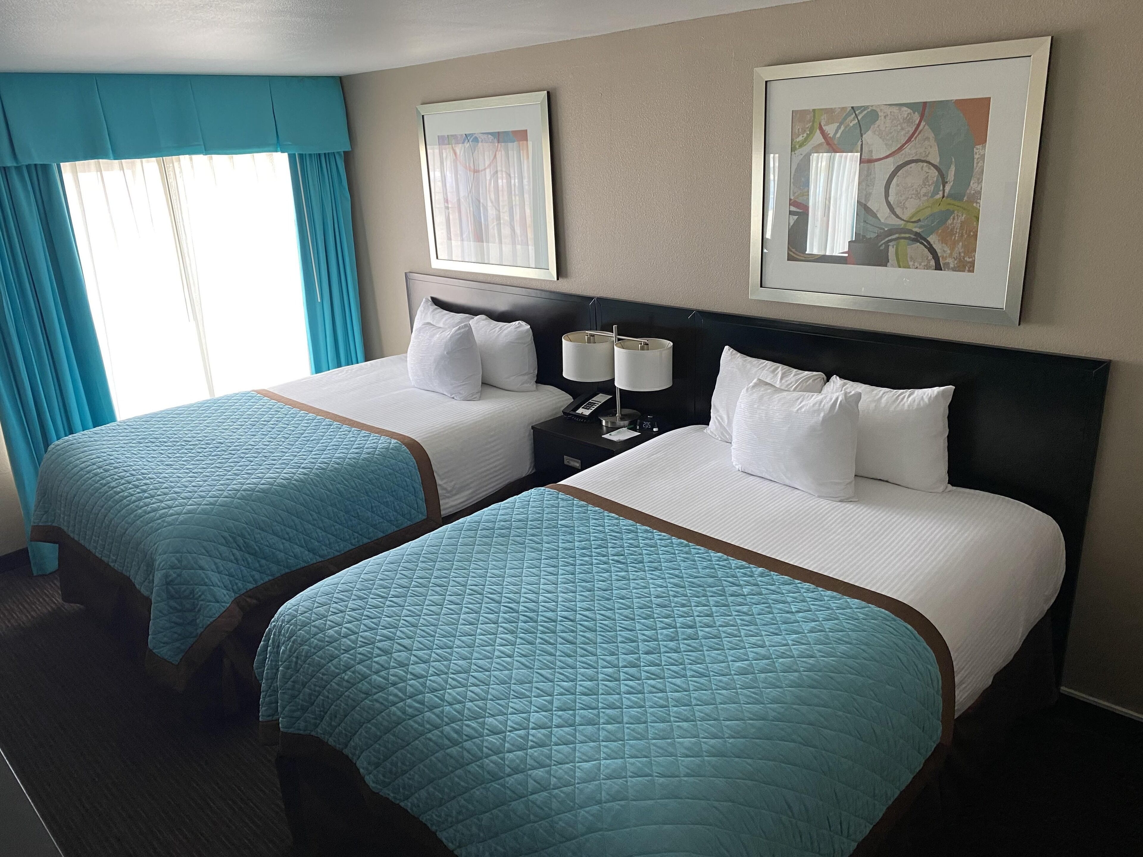 A hotel room with two beds, each with a blue quilted bedspread and white pillows. There are two framed artworks on the wall, a nightstand with a lamp and phone, and a window with blue curtains.