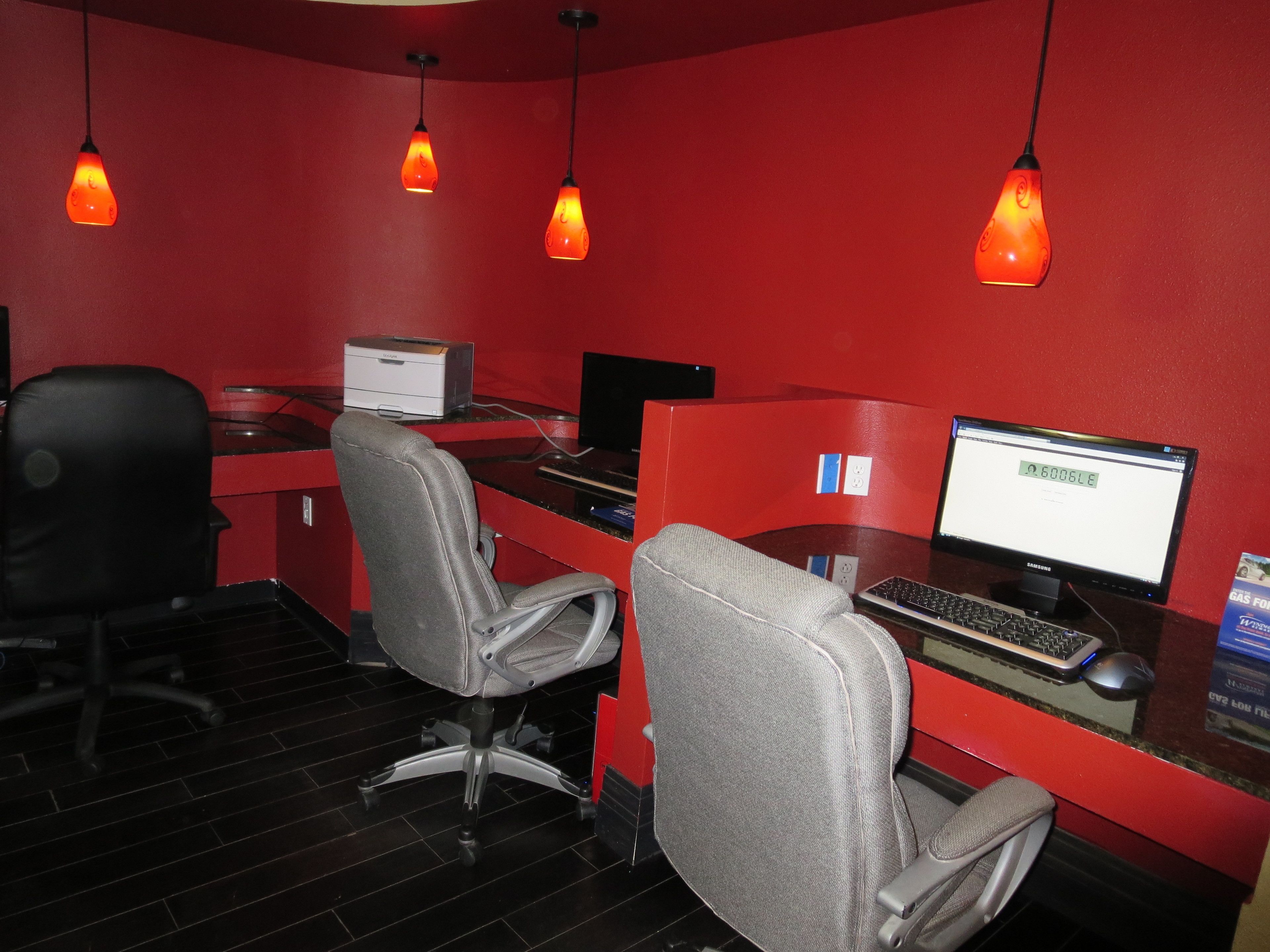 A modern office with two desks, one with a computer and keyboard, and another with a monitor displaying '6006LE'. There are two office chairs, a black chair, and a grey chair. The room has red walls and hanging orange pendant lights.