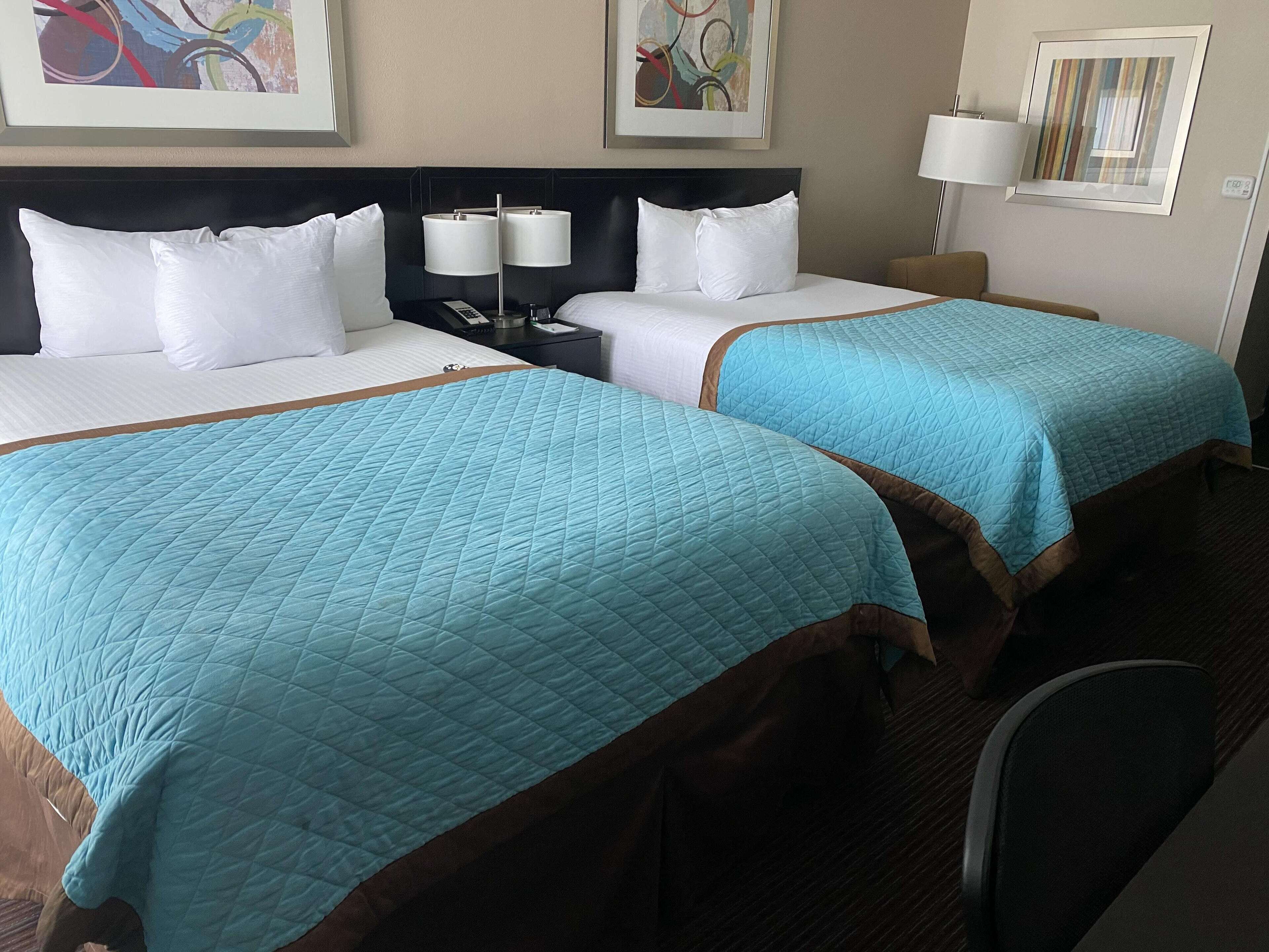 A hotel room with two beds, each with a blue quilted bedspread and white pillows. There is a nightstand with a lamp and a phone between the beds. The room has a dark wood headboard and a beige wall with abstract art.