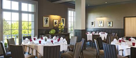 A dining area with multiple tables set for guests, large windows providing natural light, and framed pictures on the walls.
