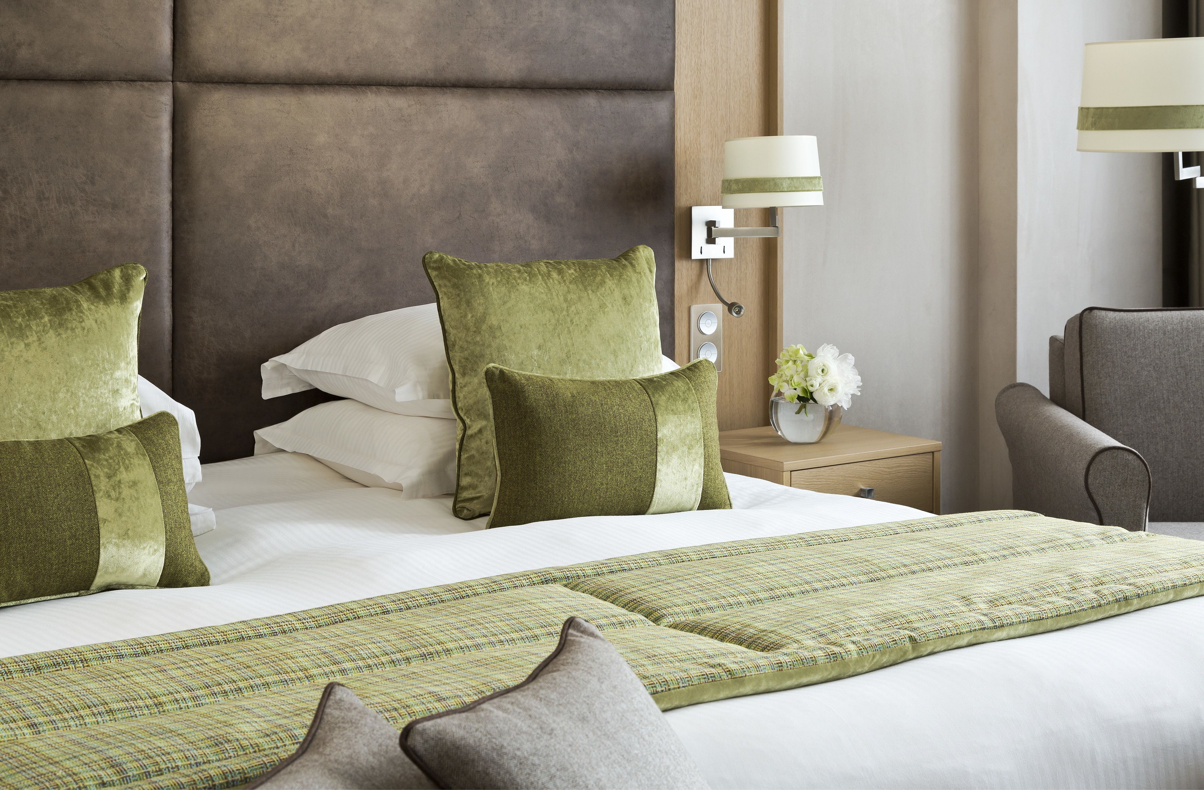 A neatly made bed with green and white pillows, a green and white checkered throw blanket, and a grey armchair.