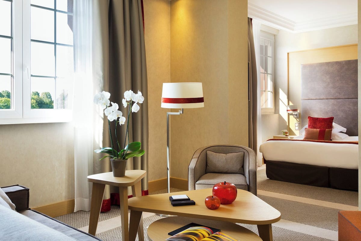 Ein Hotelzimmer mit einer Couch, einem kleinen Tisch mit Äpfeln, einem Sessel, einem Bett mit roten Kissen, einer Lampe und einem Fenster mit Vorhängen.