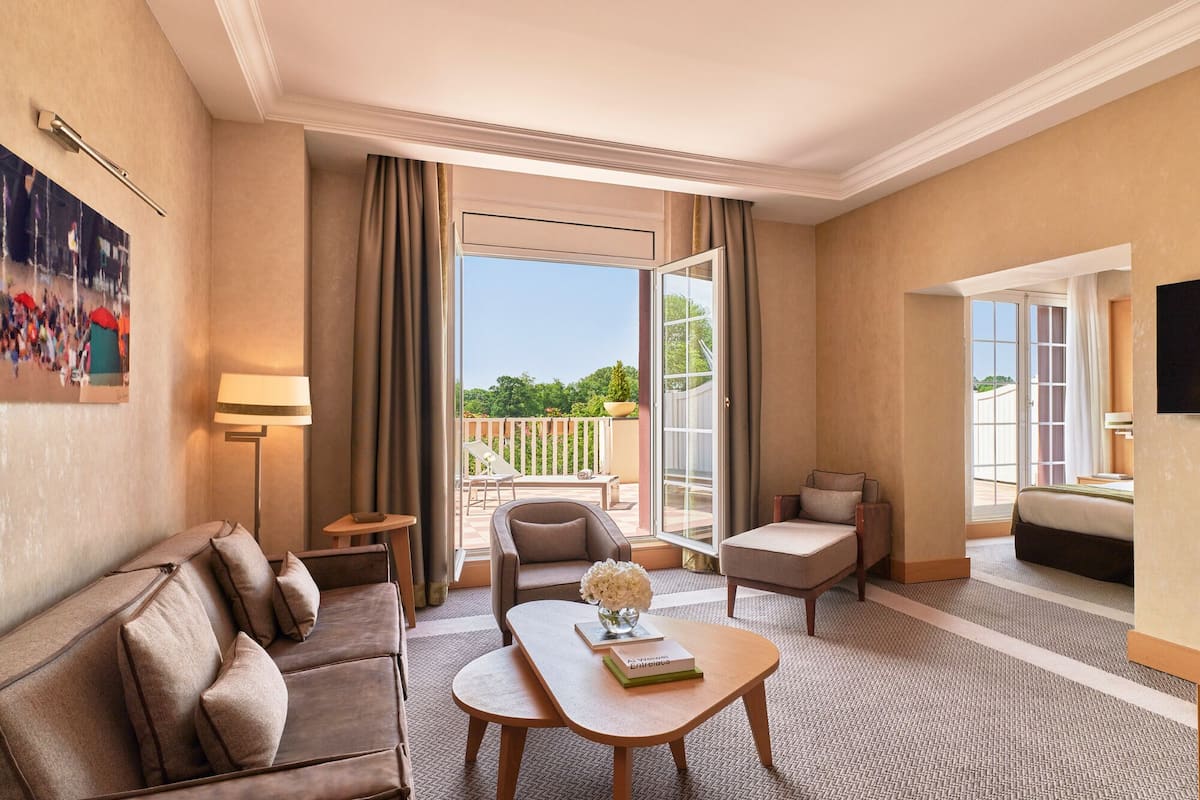 Ein Hotelzimmer mit einer Couch, einem Sessel und einem kleinen Tisch mit einem Buch und Blumen. Ein Balkon bietet Blick auf Grünflächen.