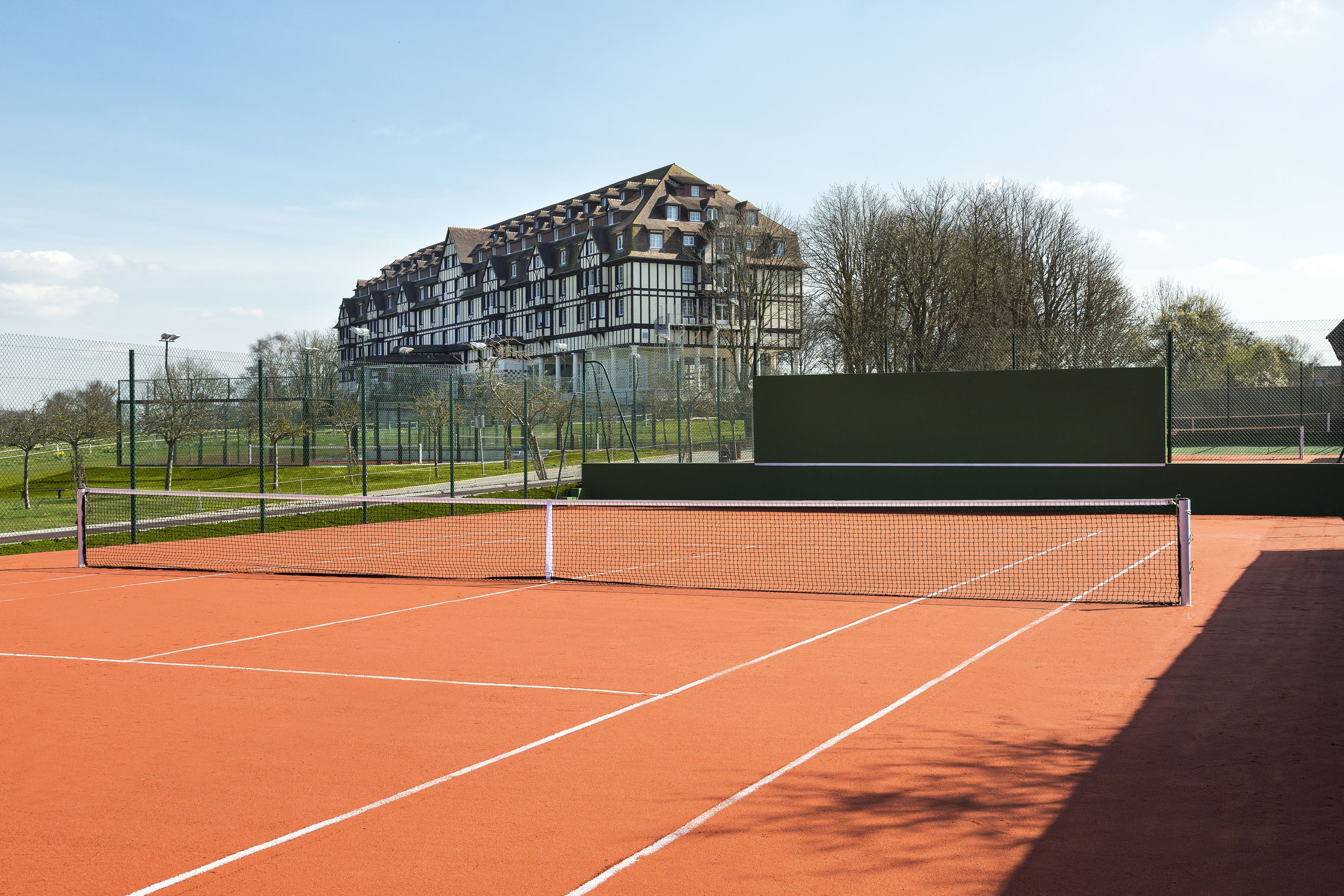 Tenniskenttä, jossa on verkko, ja jonka ympärillä on aita ja puita.