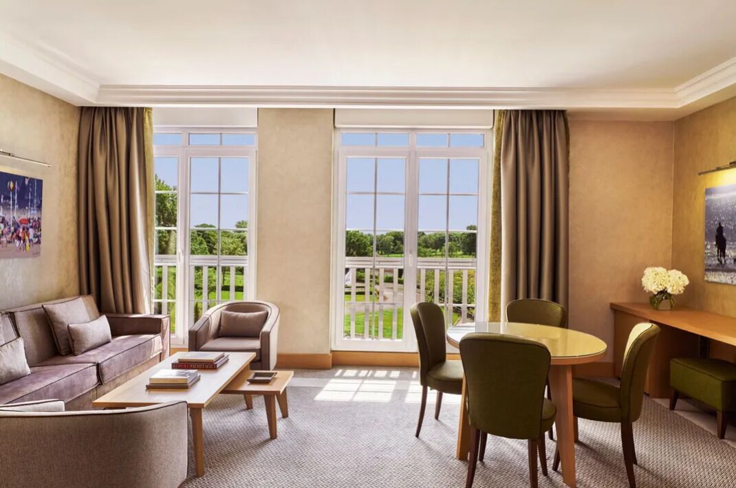 A hotel room with a large window, a sofa, a dining table with chairs, and a small side table.