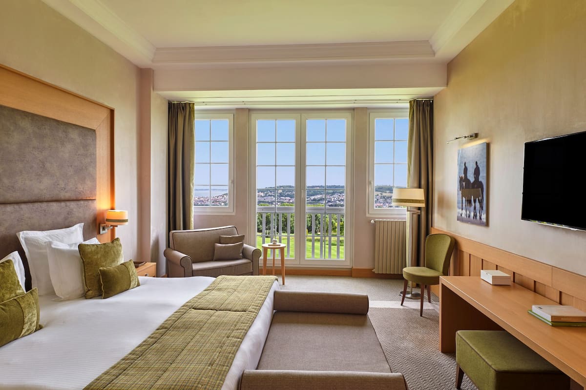 Ein Hotelzimmer mit einem großen Bett, einem Schreibtisch, einem Sessel, einem Fernseher und einem Balkon mit Blick.
