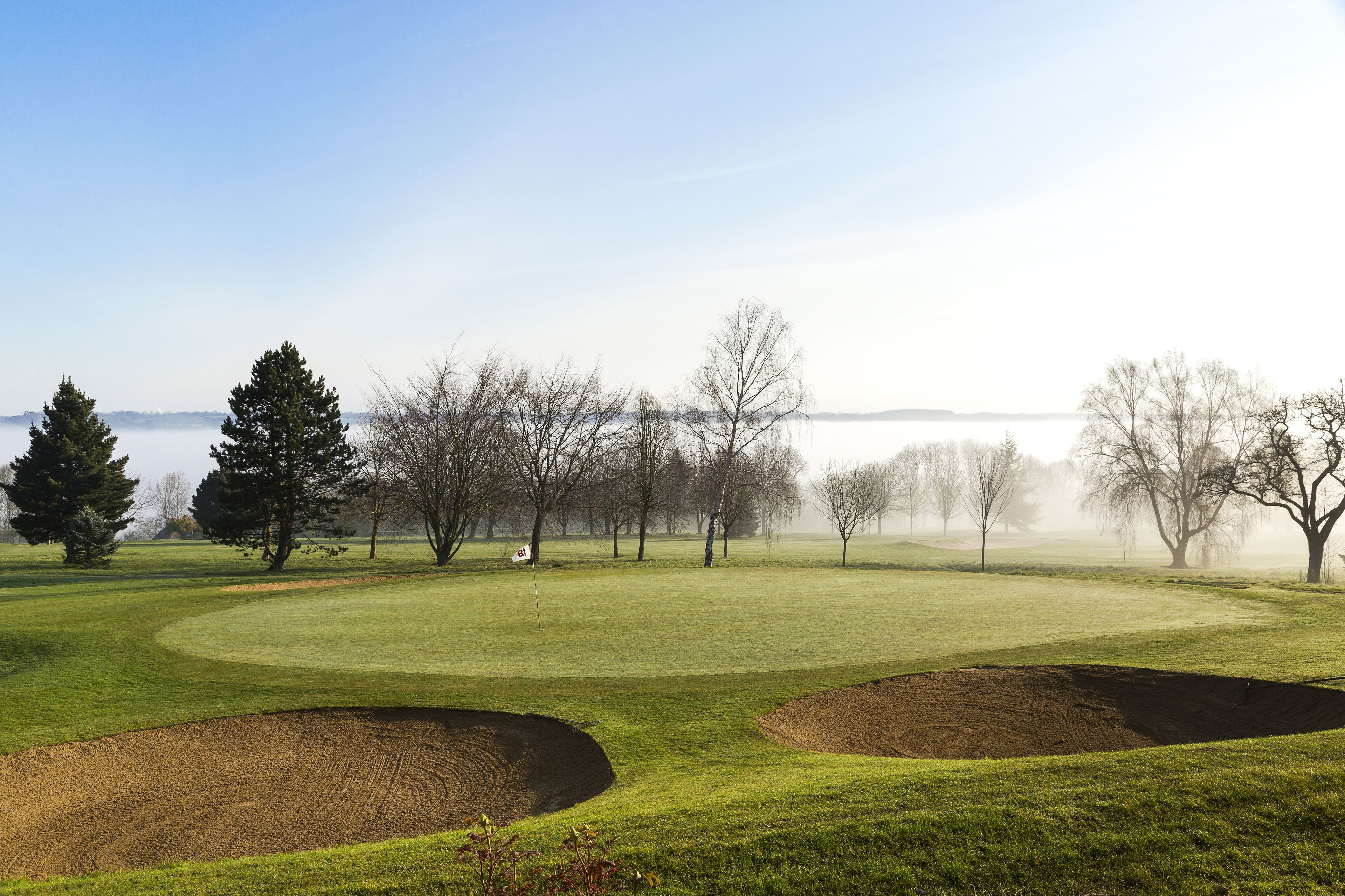 A golf course with bunkers, trees, and a body of water in the background.