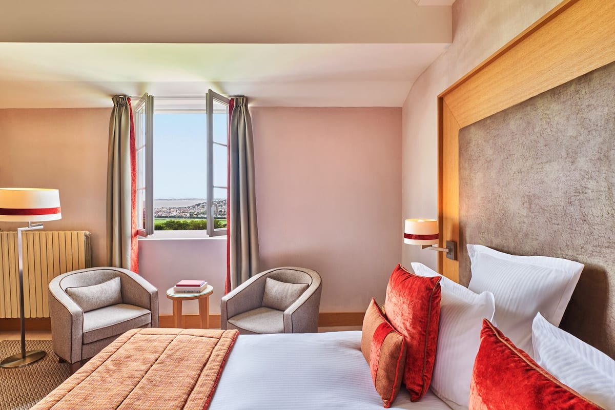 Ein Hotelzimmer mit einem Bett, zwei Sessel, einem kleinen Tisch und Blick auf die Stadt durch große Fenster.