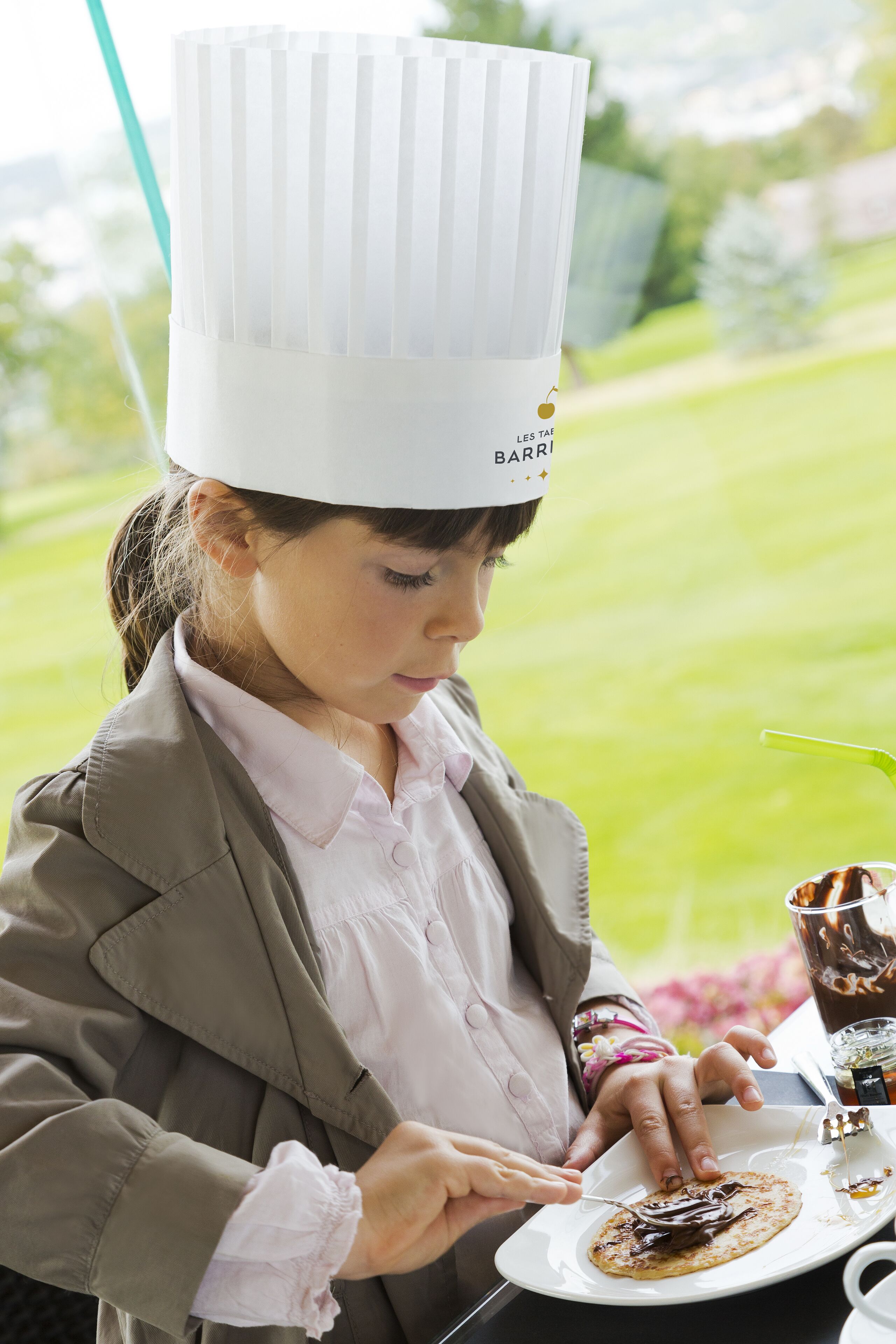 A person wearing a chef's hat and a brown jacket, holding a fork and knife, with a plate of food in front of them.
